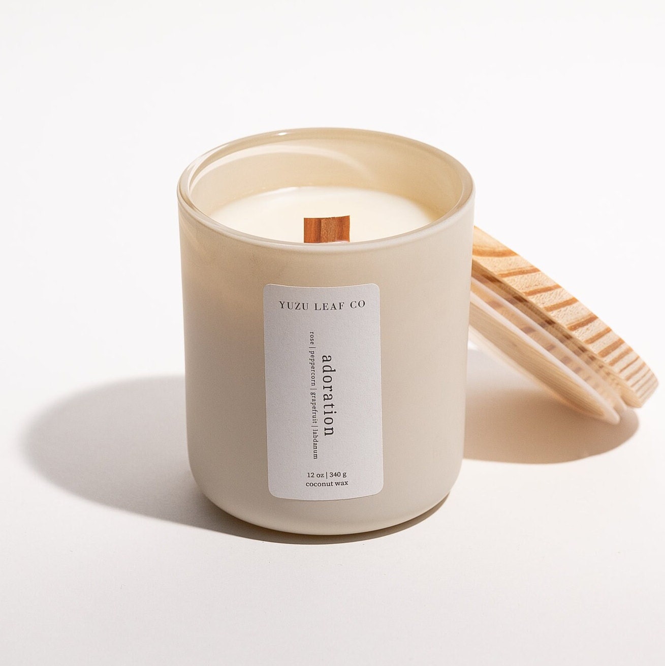 There is a large, cylindrical glass jar candle with a matte finish. The wick is made of wood and the label says "adoration: rose, peppercorn, grapefruit, labdanum." The wooden lid is leaning against the glass jar.