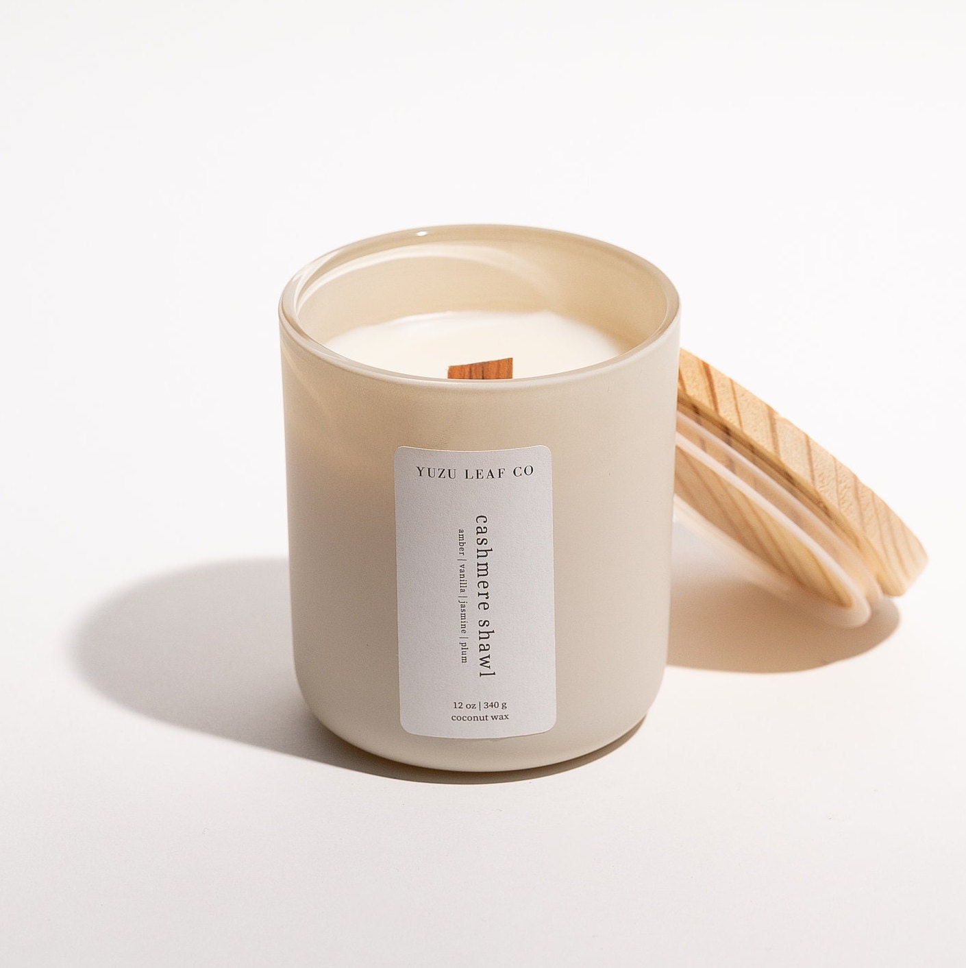 There is a large, cylindrical glass jar candle with a matte finish. The wooden lid is leaning against the glass jar, displaying the wooden wick in the middle. The vertical label says "cashmere shawl: amber, vanilla, jasmine, plum."