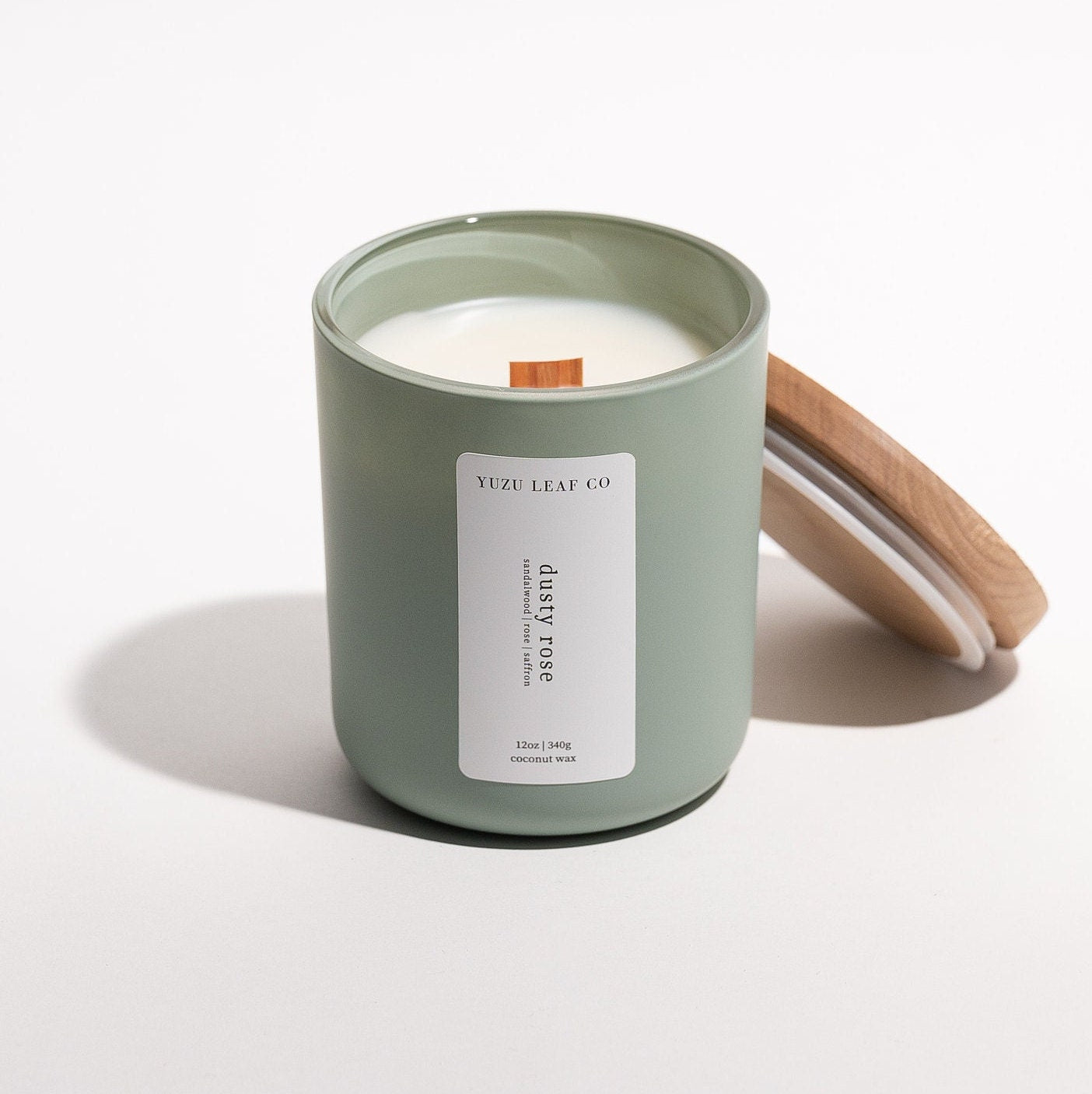 There is a large, cylindrical glass jar candle with a matte finish. The wooden lid is leaning against the glass jar, displaying the wooden wick in the middle. The vertical label says "dusty rose" with notes of sandalwood, rose, and saffron. 