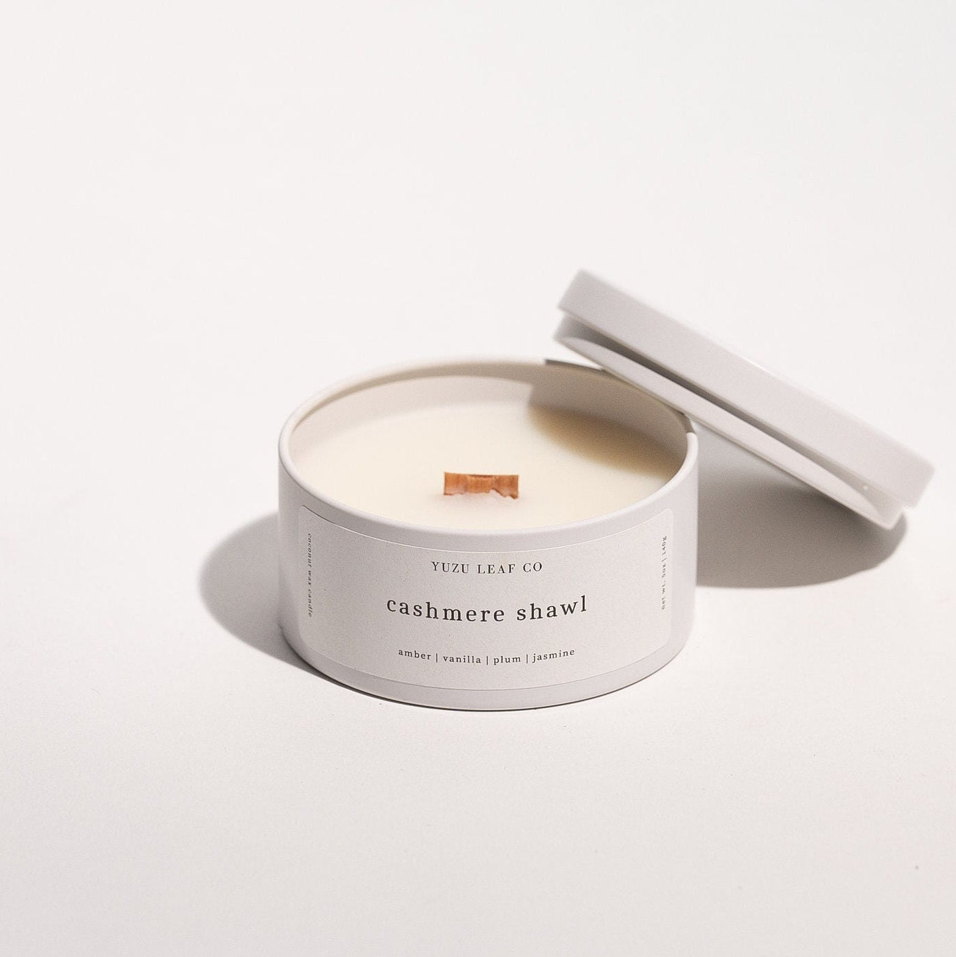 There is a candle tin that is about the size of a circular ice cream sandwich or small burger. The lid is off and leaning against the side to display the wooden wick in the middle. The candle is named "cashmere shawl" with notes of amber, vanilla, plum, and jasmine.
