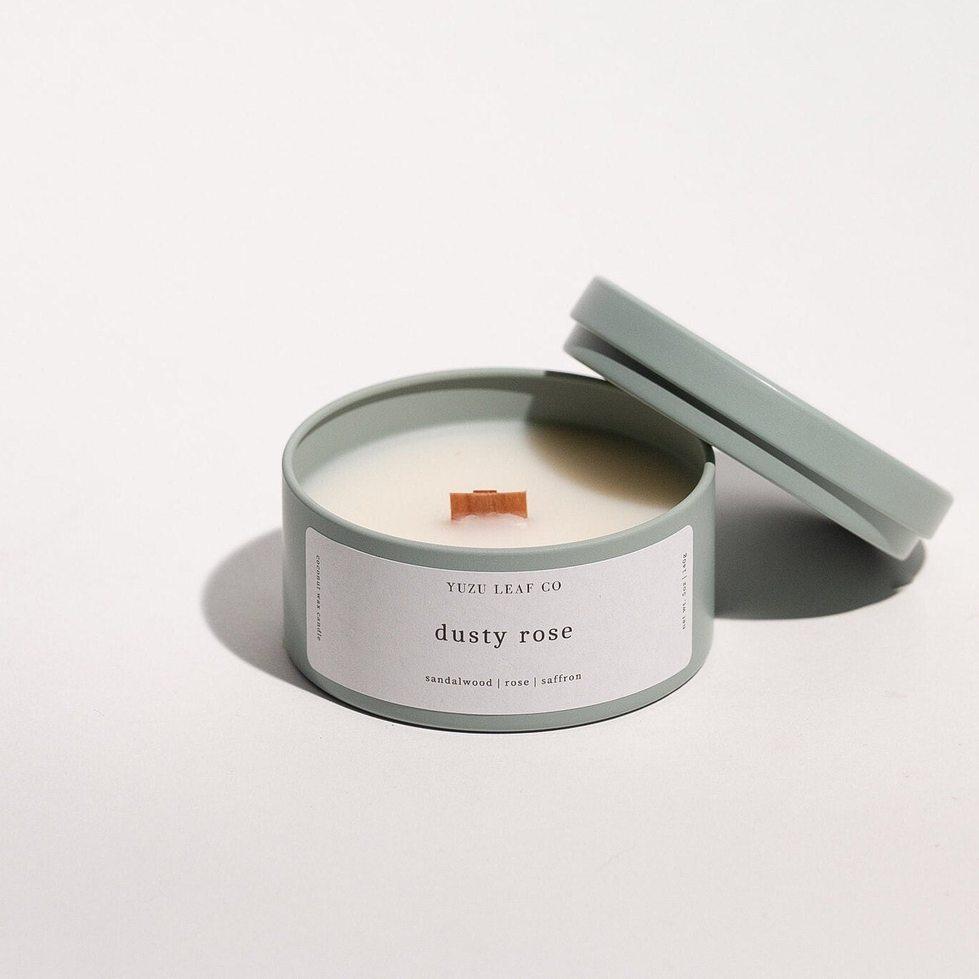 There is a candle tin that is about the size of a circular ice cream sandwich or small burger. The lid is off and leaning against the side to display the wooden wick in the middle. The candle is named "dusty rose" with notes of sandalwood, rose, and saffron.