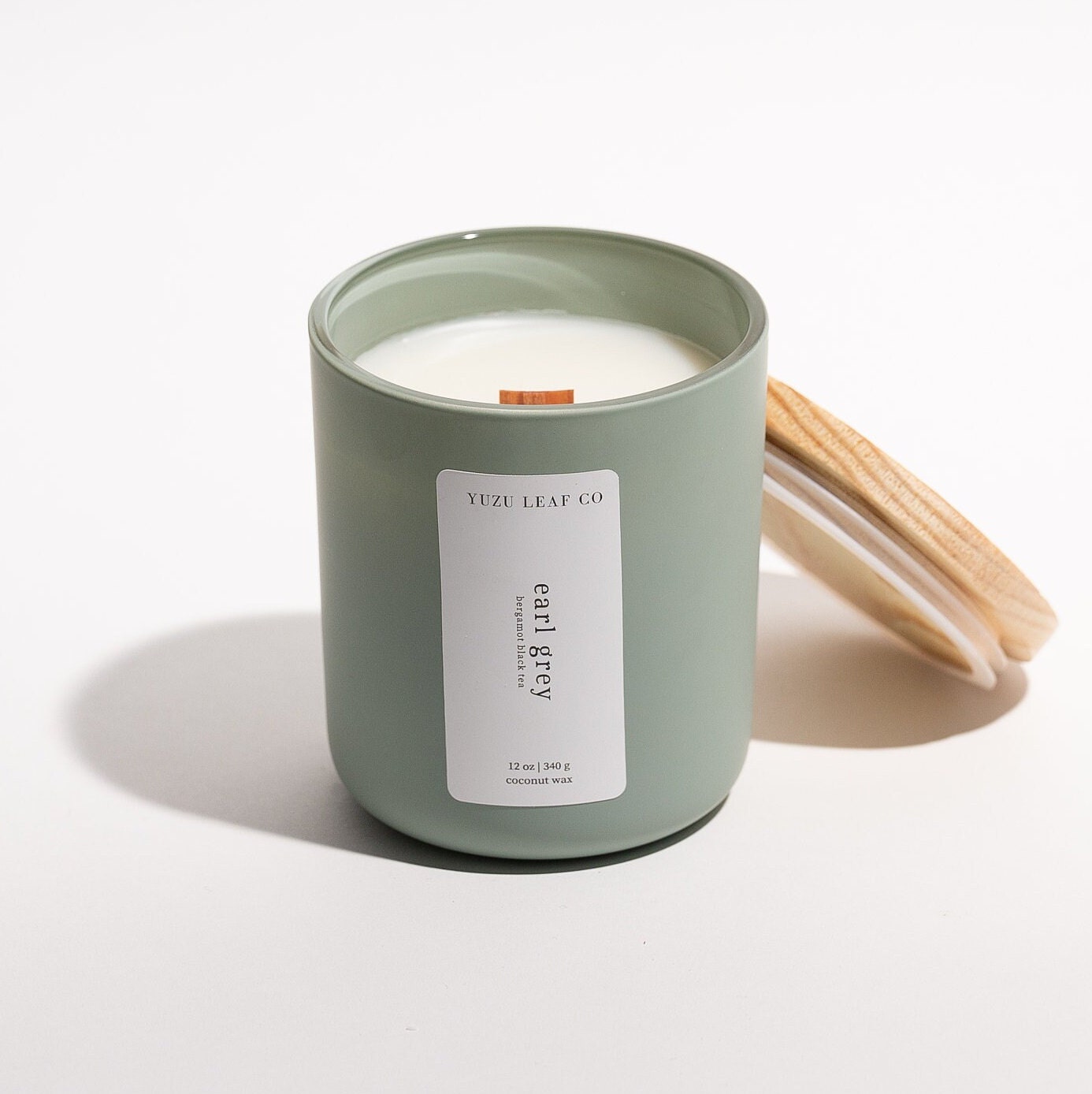 There is a large, cylindrical glass jar candle with a matte finish. The wooden lid is leaning against the glass jar, displaying the wooden wick in the middle. The vertical label says "earl grey: bergamot black tea." 