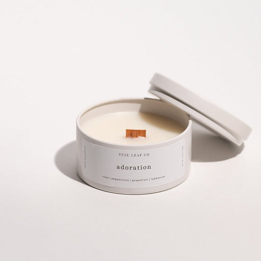 There is a candle tin that is about the size of a circular ice cream sandwich or small burger. The lid is off and leaning against the side to display the wooden wick in the middle. The candle is named "adoration" with notes of rose, peppercorn, grapefruit, and labdanum.